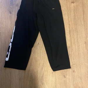 Svarta mjukisbyxor från GASP - Säljer ett par svarta mjukisbyxor från GASP med stort vitt och orange tryck längs ena benet. Byxorna har resår i midjan och en avslappnad passform, perfekta för gymmet eller chill hemma. Vid frågor kontakta mig. Priset är prutbart vid snabb och smidig affär!