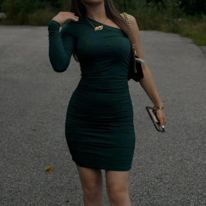 Mörkgrön one shoulder klänning - Säljer en mörkgrön, tight klänning med one shoulder-design. Klänningen är kort och har rynkade detaljer längs sidorna som framhäver figuren. Perfekt för dig som vill sticka ut med en elegant och modern look.