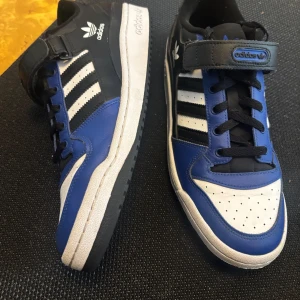 Adidas sneakers blå och svart med kardborre - Säljer ett par Adidas sneakers i blått, svart och vitt med klassiska tre ränder på sidan. Skorna har snörning och kardborreband upptill samt vit sula. Perfekta för dig som gillar sportig stil och vill ha bekväma skor till vardagen.