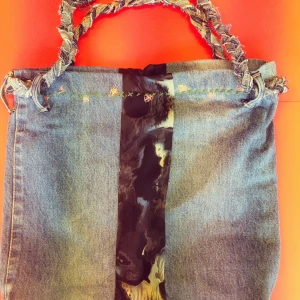 Jeansväska med broderad detalj (kan bli till påse)  - Unik handväska i jeans med flätat handtag och en svartmönstrad mittpanel. Mittpanelen gör så att du kan ändra handväskan till en "påse". (Backsidan är inte backsidan, det är bara hur den ser ut när man drar i handtaget. Den är INTE broderad i baksidan som i framsidan. Annars ser allt likadant ut