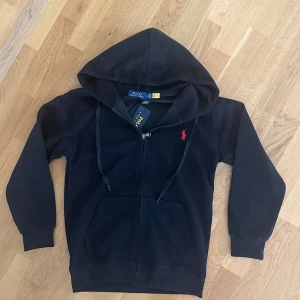 Svart ralph lauren zip hoodie - Säljer en svart hoodie från Polo Ralph Lauren kofta. Tröjan har en röd broderad logga på bröstet och två fickor framtill. Perfekt för en avslappnad stil och enkel att matcha med det mesta. Skit snygg nu till sommar. Storlek S men passar nog XS också.