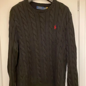 Svart stickad tröja från Ralph Lauren - Säljer en klassisk svart stickad tröja från Ralph Lauren med kabelstickat mönster. Tröjan har en rund halsringning och långa ärmar. Den ikoniska röda logotypen är broderad på bröstet. Perfekt för kyliga dagar! Priset är ej hugget i sten!