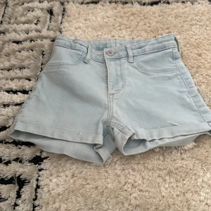 Ljusblå jeansshorts - Ljusblå jeansshorts med klassisk femficksdesign och uppvikta benslut. Hög midja och stängs med knapp och dragkedja. Perfekta för varma dagar och enkel att matcha med olika toppar.