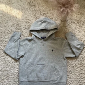 Grå hoodie från Polo Ralph Lauren - Säljer en klassisk grå hoodie från Polo Ralph Lauren med broderad logga på bröstet. Tröjan har huva, magficka och ribbade muddar vid ärmslut och nederkant. Perfekt för en avslappnad stil.