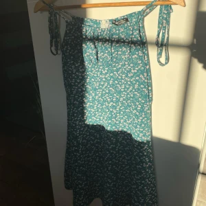 Blommig blå klänning med knytband - Söt blå klänning med små vita blommor och knytband på axlarna. Lätt och luftig modell, perfekt för varma dagar. Klänningen har en avslappnad passform och är ärmlös.