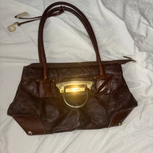 Brun handväska från Dolce & Gabbana - Säljer en brun handväska från Dolce & Gabbana i skinn med guldiga metalldetaljer och dubbla handtag. Väskan har en dekorativ metallplatta framtill och innerfoder med logotyp. Perfekt för dig som vill ha en snygg och klassisk accessoar.