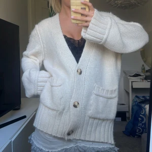 Vit stickad kofta med knappar - Mysig oversized vit stickad kofta med stora bruna knappar och två fickor framtill. Koftan har lång ärm och bred ribbad mudd vid ärmslut och nederkant. Perfekt för lager-på-lager och chill dagar.