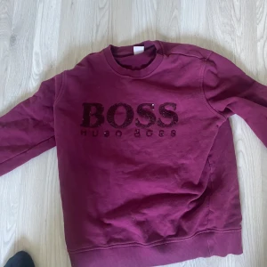 Vinröd sweatshirt från Hugo Boss - Säljer en vinröd sweatshirt från Hugo Boss med glittrig BOSS-logga framtill. Tröjan har rund halsringning och långa ärmar. Perfekt för en avslappnad och snygg stil.