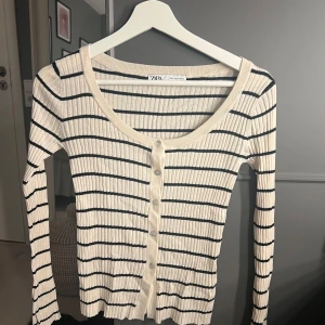 Randig ribbad kofta från Zara - Säljer en smalrandig, ribbad kofta från Zara i beige och svart. Tröjan har långa ärmar, knappar framtill och en croppad passform. Perfekt att styla över ett linne eller t-shirt.