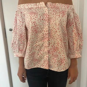 Offshoulder blus med röda prickar - Säljer en söt offshoulder blus i vitt med röda prickar och trekvartsärmar. Blusen har knappar framtill och en avslappnad passform, perfekt för vår och sommar.