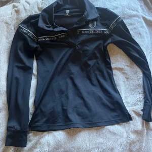 Svart långärmad pikétröja från Maya Delorez - Säljer en svart långärmad pikétröja från Maya Delorez med klassisk krage och logga på bröstet. Tröjan har diskreta logoband längs axlarna och är tillverkad i ett stretchigt material som passar perfekt till ridning. Bara använd 1 gång 