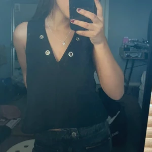 Svart croppad linnetopp med silvriga öljetter - Säljer en svart croppad linnetopp med silvriga öljetter vid halsringningen både fram och bak. Har klippt den själv.