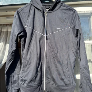 Svart jacka från Nike - Säljer en svart jacka från Nike med vit dragkedja och diskret logga på bröstet. Jackan har huva, långa ärmar och två fickor framtill. Perfekt för träning eller chill.