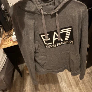 Grå hoodie från Emporio Armani EA7 - Säljer en grå hoodie från Emporio Armani EA7 med stor logga framtill. Tröjan har huva med dragsko och en stor magficka. Perfekt för dig som gillar stilrena och bekväma plagg.