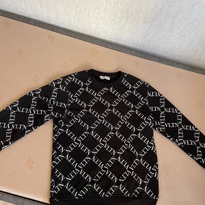 Svart sweatshirt från Valentino med VLTN-logga - Cool svart sweatshirt från Valentino med vit VLTN-logga över hela tröjan. Rund halsringning och långa ärmar. Perfekt för dig som gillar streetwear och vill sticka ut med en designertröja. Passar folk med S och M.