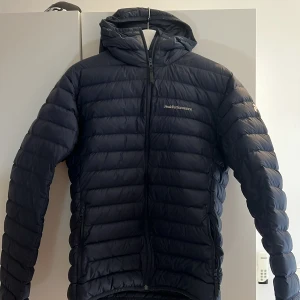 Svart dunjacka från Peak Performance - Säljer en mörkblå dunjacka från Peak Performance med huva och dragkedja. Jackan är lätt, har quiltad design och två sidofickor med dragkedja. Jackan är mycket bra skick och är i storlek L. 