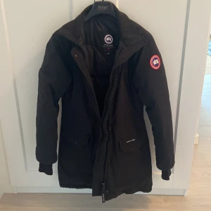 Svart dunjacka från Canada Goose - Säljer en svart lång dunjacka från Canada Goose med klassisk logga på ärmen och fickor framtill. Jackan har dragkedja och knappar, samt lång ärm med muddar. Den har också en luva som jag har valt att ta bort men den finns med.