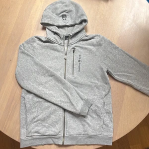 Grå hoodie från Sail Racing - Säljer en grå hoodie från Sail Racing med dragkedja framtill och bröstficka med dragkedja. Jackan har broderad logga på bröstet och huva. Perfekt för svala dagar och enkel att matcha med det mesta. Är som en S i storlek.