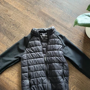 Svart hybridjacka från Everest - Svart jacka från Everest i storlek 158/164. Jackan har quiltad framsida och rygg med släta svarta ärmar. Stängs med dragkedja och har hög krage. Perfekt för kyliga dagar.