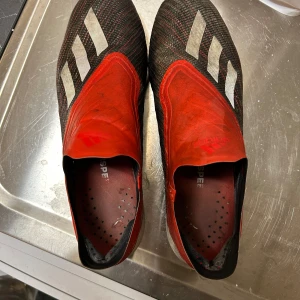 Adidas fotbollsskor  - Säljer ett par Adidas  fotbollsskor i rött och svart med vita detaljer och tre klassiska ränder på ovansidan. Skorna har FG-dobbar. Perfekta för spel på gräsplan. Storlek 41 1/2. Priset går att diskutera!