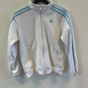 Vit Adidas track jacket med blå ränder - Vit track jacket från Adidas med ljusblå ränder längs ärmarna och dragkedja framtill. Jackan har hög krage, två fickor med dragkedja och ribbade muddar vid ärmslut och nederkant. Klassisk Adidas-logga på bröstet.