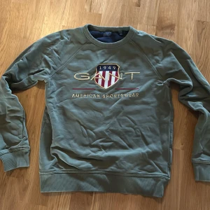 Olivgrön sweatshirt från Gant - Säljer en olivgrön sweatshirt från Gant med broderad logga och texten 'American Sportswear' framtill. Tröjan har rund halsringning, långa ärmar och ribbade muddar vid ärmslut och nederkant. 