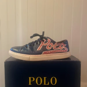 Blå sneakers från Polo Ralph Lauren - Snygga blå sneakers från Polo Ralph Lauren med stort Polo-tryck på sidan och vita sulor. Skorna har snörning och rund tå, perfekt för en sportig och avslappnad stil. Har tyvärr inte kvar originallådan, men skickar med en blå som passar bra till. Priset kan diskuteras.
