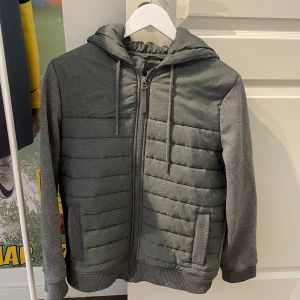 Grå quiltad hoodiejacka med dragkedja - Snygg grå hoodiejacka med quiltade partier framtill och på sidorna. Jackan har huva med snörning, dragkedja hela vägen och ribbade muddar vid ärmslut och nederkant. Perfekt för lager-på-lager under kyliga dagar.