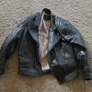 Svart skinnjacka från Halvarssons - Cool svart skinnjacka med äkta skinn från Halvarssons. Jackan har flera dragkedjeförsedda fickor, bältesdetaljer i midjan och klassisk bikerstil. Perfekt för dig som gillar en tuff look.