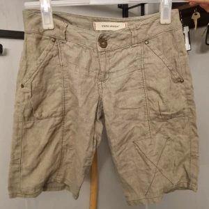 Beige shorts från Vero Moda - Beiga shorts från Vero Moda med blommigt tryck på bakfickan och dekorativa knappar. Modellen har flera fickor och är tillverkad i ett linne material. Perfekta för varma dagar.