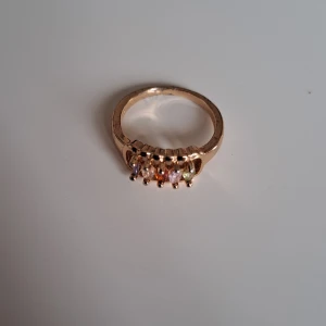 Guldfärgad ring med färgade stenar - Snygg guldfärgad ring med fem gnistrande stenar i olika färger – lila, rosa, orange och grön. Perfekt för dig som vill ha en färgklick på handen och gillar unika smycken.