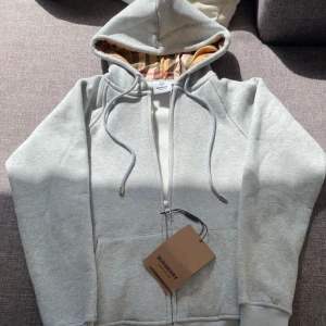Ljusgrå hoodie med dragkedja från Burberry - Hej! Säljer en burberry kofta som är HELT ny och jag säljer den på grund av att den var för liten. Om ni har frågar så skriv. Pris kan diskuteras