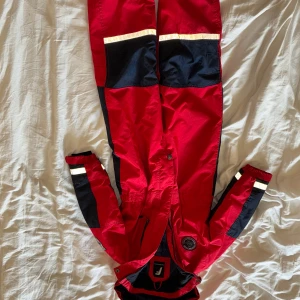 Röd och svart overall från Jofama för barn  XS storlek  - Säljer en röd och svart overall från Jofama med huva och reflexdetaljer. Overallen har dragkedja framtill, svarta förstärkningar på knän och armbågar samt vita reflexband på ärmar och ben. Perfekt för kalla dagar utomhus.