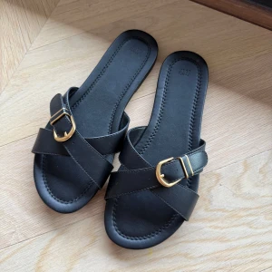 Svarta sandaler - Säljer ett par svarta sandaler från H&M med korsade remmar och snyggt guldfärgat spänne. Platt sula och öppen tå. Storlek 38, aldrig används