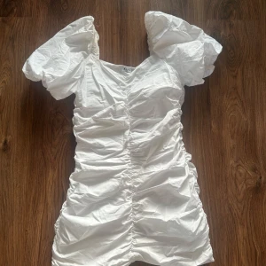 Vit kortklänning med puffärmar och rynkade detaljer - Supersöt vit klänning med puffiga kortärmar och rynk längs hela framsidan. Klänningen har en feminin v-ringning och figurnära passform som framhäver siluetten. Perfekt för studenten och sommarens alla tillfällen! 💓(den är liten i storleken)