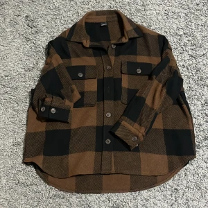 Brun och svart rutig overshirt - Säljer en brun och svart rutig overshirt från Gina tricot. Skjortan har stora rutor, två bröstfickor med knappar och klassisk krage. Perfekt att slänga över en t-shirt för en avslappnad look.