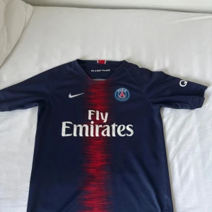 PSG fotbollströja Mbappé #7 - Mörkblå Paris Saint-Germain fotbollströja med röd vertikal rand, Nike-logga och Fly Emirates sponsortryck. Mbappé och nummer 7 på ryggen. Autentisk märkning och texten 'ICI C'EST PARIS' i nacken. Perfekt för fans av PSG och Mbappé.