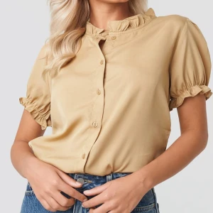 Kortärmad blus med fransig krage från NA-KD - Beige blusen från NA-KD, har en hög och fransig krage, knappar ner längs framsidan, ett elastiskt band på ärmarna och en fransig detalj på ärmarna. Sparsamt använd. (Den svarta är såld).