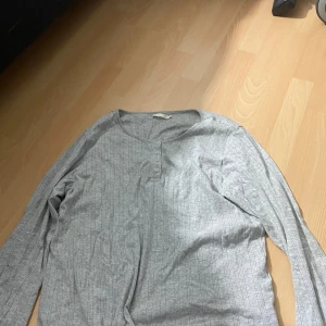 Grå ribbad långärmad tröja från 157 - Säljer en grå långärmad tröja från 157 med diskret ribbat mönster och knappar vid halsringningen. Perfekt basplagg med enkel och stilren design.