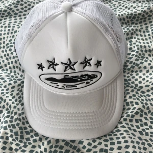 Vit truckerkeps från Corteiz - Säljer en vit truckerkeps från Corteiz med svart broderad logga och stjärnor framtill. Kepsen har mesh baktill för extra ventilation och justerbar passform. Perfekt för dig som gillar streetwear och snygga accessoarer.