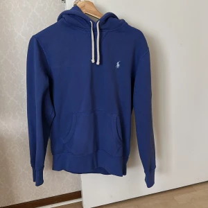 Blå hoodie från Ralph Lauren - Säljer en blå hoodie från Ralph Lauren med vit dragsko och klassisk logga broderad på bröstet. Tröjan har känguruficka och långa ärmar, perfekt för en avslappnad stil.