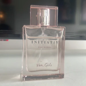 Initiative For Women parfym från Van Gils - Elegant parfymflaska i transparent glas med svagt rosa ton och stilren design. Flaskan har raka linjer och en fyrkantig form, med texten 'Initiative For Women' och 'Van Gils' på framsidan. Perfekt för dig som gillar klassiska och feminina dofter.