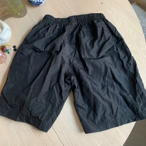 Svarta shorts från Stone Island - Säljer ett par svarta shorts från Stone Island med broderad logga på benet. Shortsen har elastisk midja med snörning och dragkedjefickor samt bakficka . Perfekta för sommaren och har en clean, stilren look.