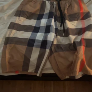 Rutig badshorts från Burberry - Snygga badshorts från Burberry med klassiskt rutigt mönster i beige, svart, vitt och rött. Shortsen har dragsko i midjan och sidofickor. Perfekta för sommaren och strandhäng.
