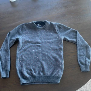 Grå stickad tröja med små röda,svarta och vita prickar - Mysig grå stickad tröja från bondelid med små röda,svarta och vita prickar över hela framsidan. Tröjan har rund halsringning, långa ärmar och ribbade muddar vid ärmslut och nederkant. Perfekt för kyligare dagar.