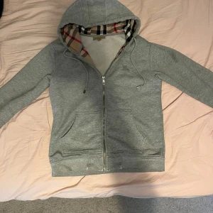 Burberry hoodie - Säljer en grå hoodie från Burberry med klassiskt rutigt foder i huvan och dragkedja framtill. Jackan har två fickor, långa ärmar och diskret broderad logga på bröstet. Perfekt för en stilren och avslappnad look.