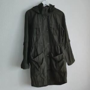 Olivgrön parkajacka från Sisters - Säljer en olivgrön parkajacka från Sisters i storlek 38. Jackan har huva, dragsko i midjan och stora fickor framtill. Stängs med knappar och dragkedja. Perfekt för kyligare dagar och enkel att styla till olika outfits.