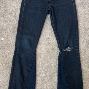 Mörkblå jeansbyxor från Levi's - Mörkblå jeansbyxor från Levi's med klassisk femficksdesign och knappgylf. Byxorna har en liten slitning på ena benet. Byxorna är i storlek W26/L30