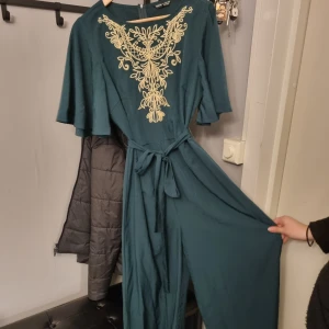 Petrolblå byxdress med broderi och knytband - Säljer en petrolblå byxdress med vackert broderi i beige framtill. Vida ärmar och knytband i midjan ger en snygg siluett. Perfekt för dig som vill sticka ut med stilrena detaljer och bekväm passform.