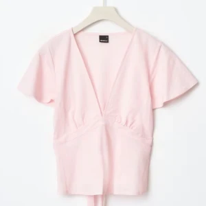 Ljusrosa omlottopp med v-ringning - Söt ljusrosa omlottopp med djup v-ringning och korta ärmar. Toppen har en feminin passform och knytning i midjan för extra detalj. Perfekt för vår och sommar!